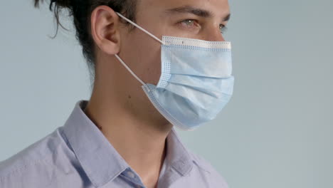 Male-with-a-Man-Bun-and-Smoldering-Eyes-Wearing-a-Button-Down-Shirt-puts-on-a-COVID-19---Coronavirus-Surgical-Mask-on-a-Grey-Background,-Side-Angle-Static-Shot