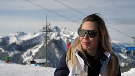 Medium-closeup-of-a-Caucasian-woman-with-brown-hair-wearing-sunglasses-and-drinking-beer-at-a-skiing-resort-with-skiing-cable-cars-and-snow-covered-mountains-in-the-background