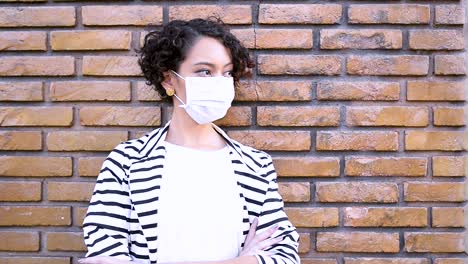Depressed-young-brunette-woman-with-coronavirus-mask-leaning-against-the-brick-wall-crosses-her-arms