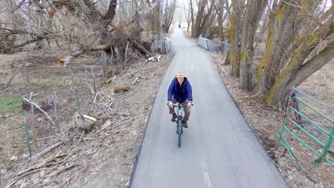 Mature,-senior-man-cycling-along-a-path-between-trees-in-the-countryside---aerial-view