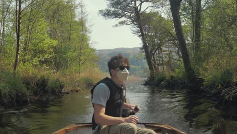 Young-man-driving-outboard-lake-boat-wearing-mask-surrounded-by-nature