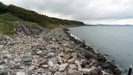 Lush-rocky-North-Wales-hillside-shoreline-ocean-boulders-aerial-view-low-angle-push-in