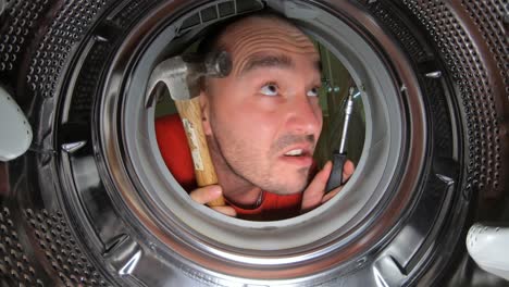 A-Man-Holding-Hammer-And-Screw-Driver-Looking-What-To-Fix-Inside-The-Washing-Machine---Closeup-Shot