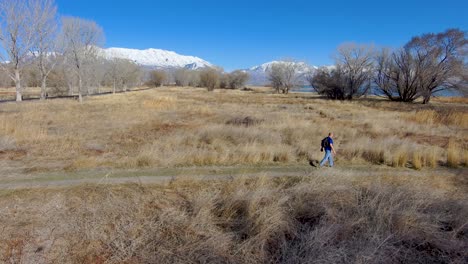 Mature-man-hiking-with-a-backpack-along-a-nature-trail-with-snow-capped-mountains-in-the-distance---aerial-view