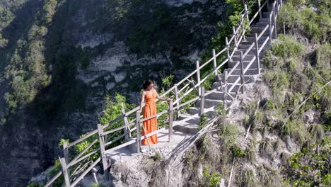 Woman-admiring-Diamond-Beach-in-Nusa-Penida-island-Indonesia-from-above-the-cliffside-staircase,-Aerial-dolly-out-reveal-shot