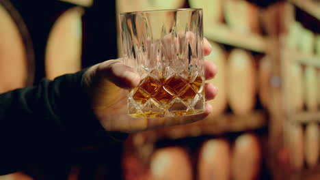 Mirando-Cuidadosamente-El-Bourbon--Mano-En-Cámara-Lenta-Girando-Un-Vaso-De-Whisky-Bourbon-Dentro-De-Una-Casa-Quieta-Con-Barriles-De-Whisky-En-Segundo-Plano