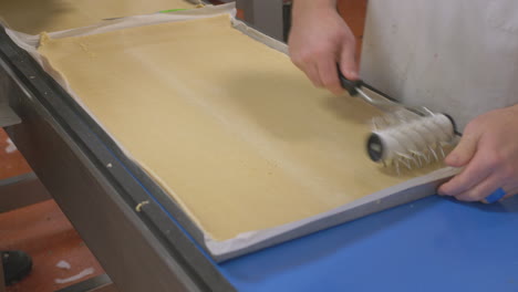 Close-up-of-a-worker-using-a-roller-docker-to-aerate-on-a-large-sheet-of-pastry