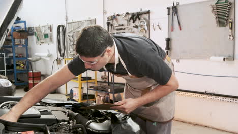 Latin-adult-car-mechanic-using-safety-glasses-checks-the-oil-level-in-the-car-engine,-in-a-repair-shop