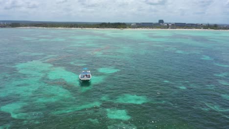 Single-motor-boat-anchored-in-turquoise-ocean-sea-water-by-Juanillo-beach,-Cap-Cana,-Dominican-Republic,-above-circle-aerial
