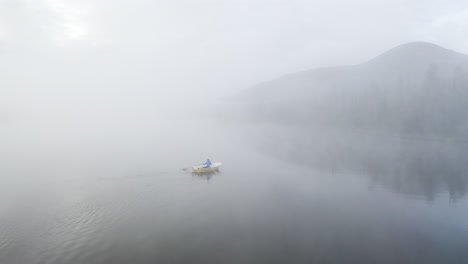 Flying-above-a-lake-next-to-a-small-boat-with-a-person-rowing-on-a-misty-morning-with-mountain-in-the-background