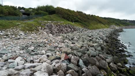 Lush-rocky-North-Wales-hillside-shoreline-ocean-boulders-aerial-view-low-angle-reverse