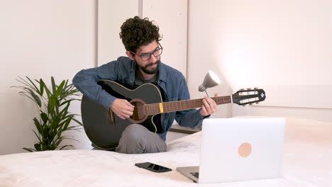Man-sits-at-home-in-bedroom-in-front-of-laptop-attending-online-guitar-lesson
