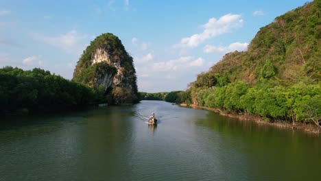 aerial-drone-flying-tracking-a-thai-longtail-boat-down-river-with-large-green-limestone-rocks-and-mangrove-forests-in-the-background-on-a-sunset-afternoon-in-Krabi-Town-Thailand