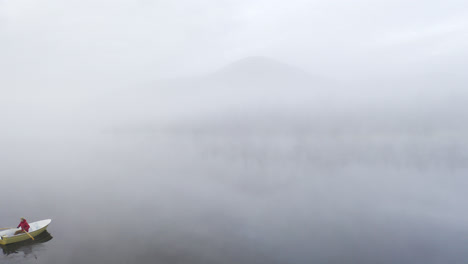 Persona-En-Un-Bote-De-Remos-En-Un-Lago-Con-Un-Paisaje-De-Niebla-Surrealista-De-Ensueño-Con-Una-Montaña-En-La-Parte-De-Atrás