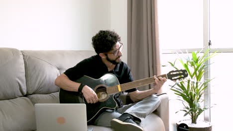 Bearded-Hispanic-man-playing-acoustic-guitar-and-singing-on-sofa-in-front-of-laptop