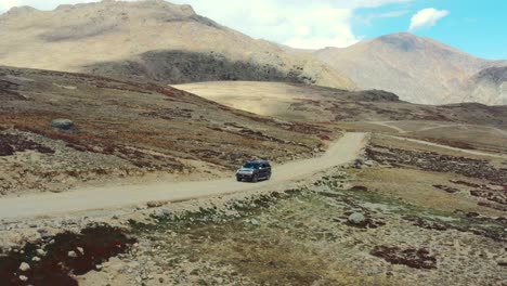 Aerial-drone-of-a-red-suv-driving-up-a-hill-slowly-on-a-dirt-road-path-in-the-high-altitude-alpine-plain-and-mountains-of-Deosai-National-Park-located-between-Skardu-and-Astore-Valley-in-Pakistan