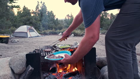 Man-Cooking-on-Campfire-at-Campsite,-Surrounded-by-Pine-Trees-and-Tents