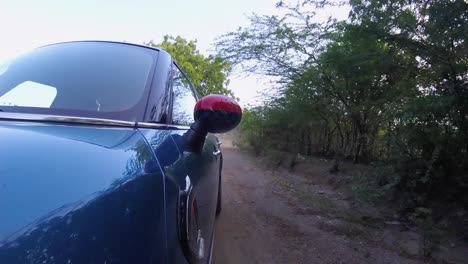 Blue-hatchback-car-moving-on-a-dusty-road,-with-red-rear-view-mirror-at-high-speed