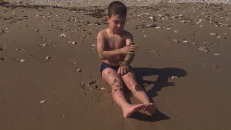 Caucasian-toddler-boy-in-a-swimsuit,-playing-with-sand,-in-sandy-beach-of-Varkiza,-Attica,-Greece