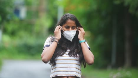Pretty-young-dark-skinned-smiling-Indian-girl-wearing-an-N95-mask-for-coronavirus-in-monsoon-rain-green-background-slow-motion
