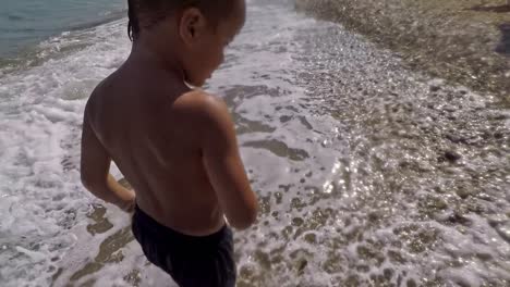 Follow-shot-of-caucasian-kid,-walking-at-the-seashore-at-Kalamata-beach,-waves-hitting-his-feet