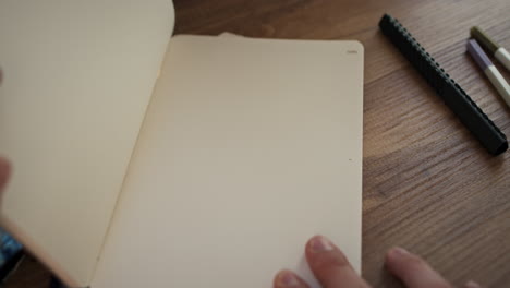 Abriendo-Un-Cuaderno-De-Trabajo-Negro-Bajo-La-Luz-Del-Sol-Encima-De-Un-Acogedor-Escritorio-De-Madera-Con-Bolígrafos