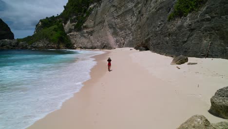 young-asian-female-model-walking-gracefully-on-a-white-sand-beach-with-the-waves-crashing-at-Nusa-Penida-Diamond-Beach-in-Bali-Indonesia-on-a-sunny-tropical-day