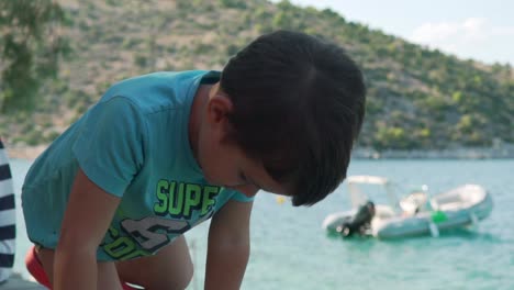 Close-up-of-caucasian-boy,-posing-at-the-camera-,sitting-on-a-ledge-besides-the-sea-of-Fragolimano,-Greece-120fps
