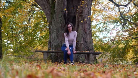 Portrait-of-Typical-Teenage-Girl-on-Bench-Under-Old-Tree-at-Park-on-Autumn-Day