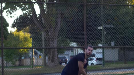 Male-with-Beard-and-Man-Bun-in-a-Park-serves-a-Tennis-Ball-with-Chain-Fence-in-Background