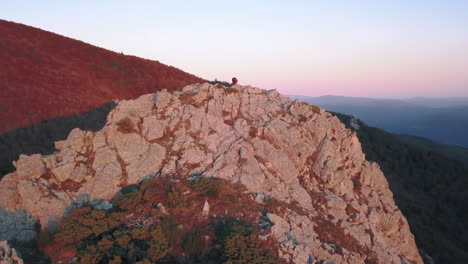 Zoom-Out-drone-shot-of-a-woman-doing-yoga-on-top-of-a-mountain-with-the-first-lights-of-sunrise