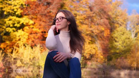 Young-Woman-in-Glasses-Sitting-on-Park-With-Trees-in-Autumn-Colors-on-Sunny-Day