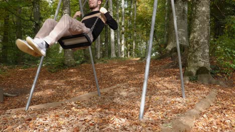 Cheerful-smiling-man-sitting-on-swing-and-swaying-during-beautiful-sunny-autumn-day-in-park