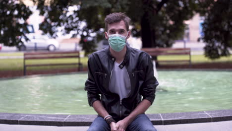 A-young-man-with-a-fashionable-hairstyle,-a-black-leatherette-jacket-and-a-white-t-shirt,-sitting-on-a-fountain-in-a-park,-removing-a-green-protective-Covid-19-mask-from-his-face,-static-4k-shot