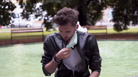 A-young-man-with-a-fashionable-hairstyle-wearing-a-black-leatherette-jacket,-white-t-shirt,-sitting-on-a-fountain-in-a-park,-removing-a-green-protective-Covid-19-mask-from-his-face,-close-up-4k-shot