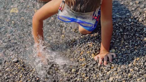 Close-up-of-caucasian-kid-digging-pebbles-at-the-sea,-trying-to-create-a-ditch-slow-motion