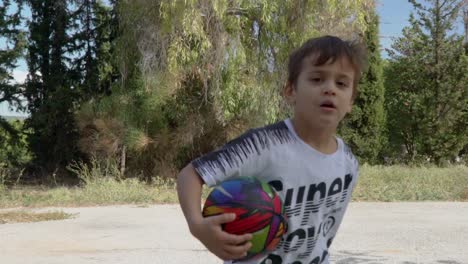 Caucasian-toddler-boy-running-around-a-small-basket-ball-outdoors,-trees-in-the-background-120fps