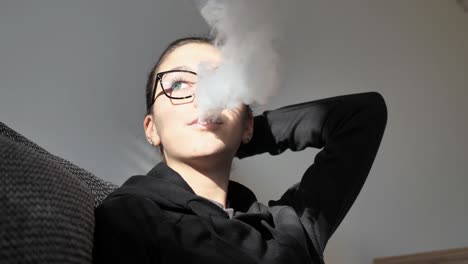 Slow-Motion-of-Young-Woman-Smoking-Electronic-Cigarette,-Releasing-Smoke-in-Room-With-Sunlight-on-Her-Face