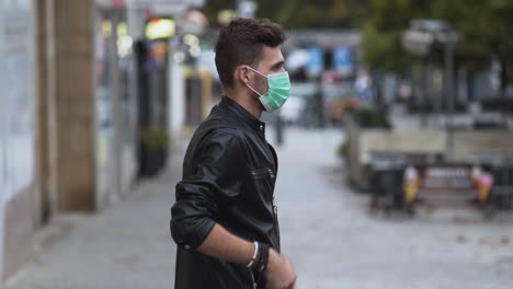 A-cool-young-man-in-a-leatherette-jacket-with-a-modern-hairstyle-on-a-busy-street-and-puts-on-a-green-protective-Covid-19-facemask,-then-putting-his-hands-in-his-pockets,-sideways-to-the-camera,-4k