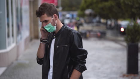 A-young-man-with-a-cool-hairstyle-wearing-a-black-jacket,-a-white-t-shirt-and-a-green-protective-Covid-19-facemask,-standing-on-a-busy-street,-picking-up-his-phone-and-calling,-static-close-up-4k