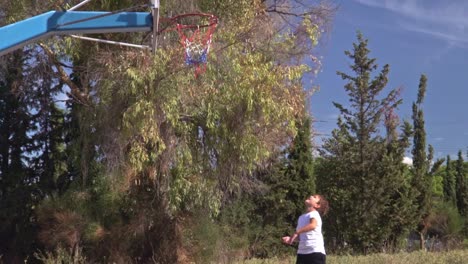 Caucasian-kid-plays-basketball-hoop-,-at-alley-basketball-court,-beautiful-trees-in-the-background