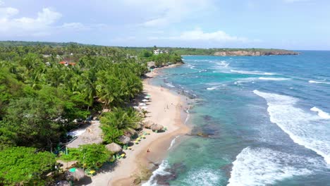 Aerial-shot-over-the-najayo-beach-with-a-view-of-thatched-houses-on-the-shore-of-the-beach-on-a-clear-afternoon,-breeze-hitting-the-palms,-sea-with-waves