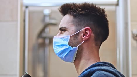 Handsome-man-blow-dry-his-hair-in-the-bathroom-wearing-protective-medical-mask