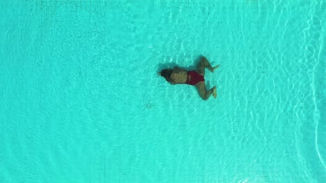 Woman-in-red-swimsuit-doing-underwater-breaststroke-on-Perenenan-Beach-Bali-swimming-pool,-Aerial-top-view-left-pan