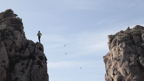A-man-checks-the-height-of-a-cliff-before-jumping,-in-the-background-seagulls-flying,-Cala-Llonga,-Ibiza,-Spain