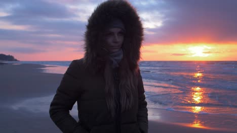 Young-woman-in-winter-clothes-walks-along-the-sandy-shore-of-the-Baltic-sea-beach-at-romantic-sunset,-medium-shot-pan-left
