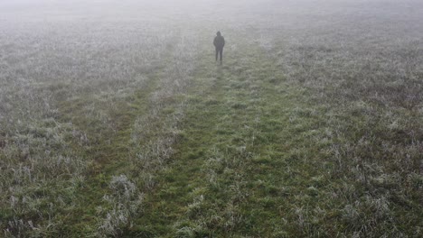 Aerial-View-of-Single-Man-Walking-in-a-Fog-on-Meadow-at-Autumn-Morning,-Tracking-Drone-Shot