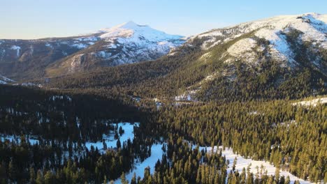 Drone-birdseye-footage-over-ski-slopes-and-surrounding-alpine-forest-on-a-clear-and-sunny-winter's-day,-moving-forward-towards-snowcapped-mountains-in-Lake-Tahoe,-USA