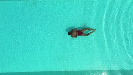 Woman-in-red-swimsuit-performing-underwater-breaststroke-on-Perenenan-Beach-Bali-pool,-Aerial-top-view-lift-shot
