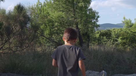 Slow-motion-shot,-back-view-of-caucasian-kid-walking-at-forest-of-Parnitha,-peaks-of-mountain-in-the-background